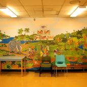 Giraffes roam through a jungle oasis of grassland and waterfalls on a large mural inside the school's front entrance. Community members gather in the foreground, dressed in vibrant prints and head coverings. Grass huts stand in the shade of tall palm trees in the distance. The mural represents St. Pat's Africentric leanings.